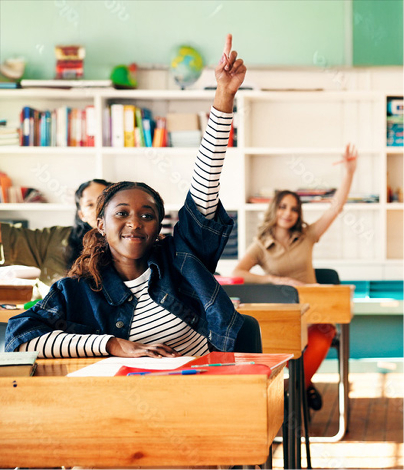 Students seated at desks in a classroom, raising hands to participate in a lesson.
